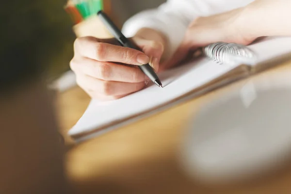 Close up of female hands writing in spiral notepad placed on wooden desktop with blurry items. Education concept — Stock Photo, Image