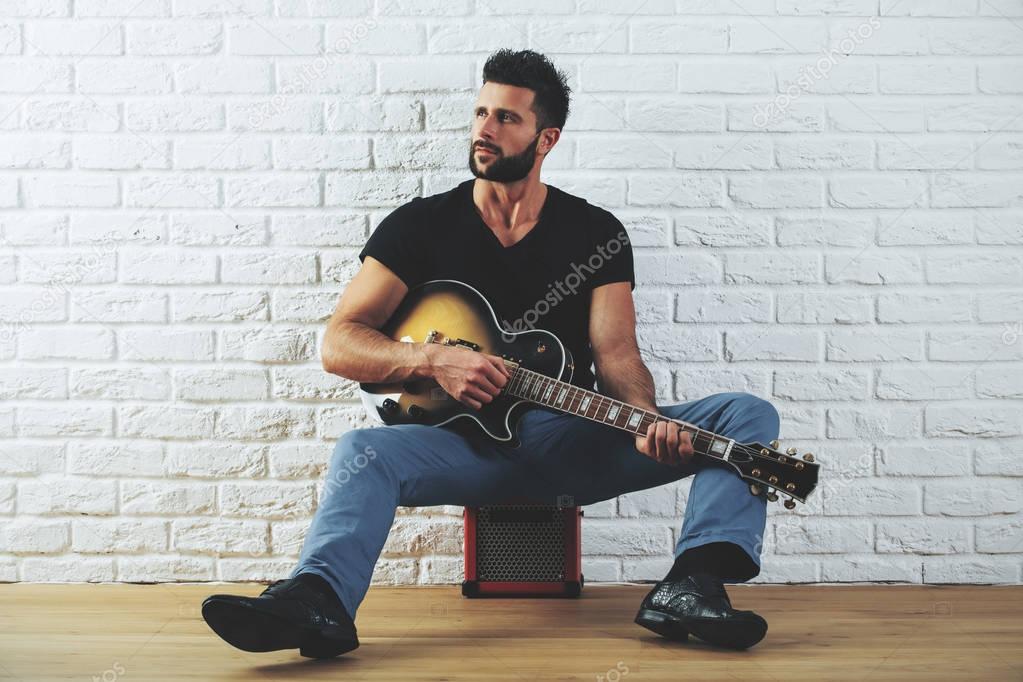 Handsome european man in casual clothing playing the guitar while standing in interior with white brick wall and wooden floor. Art concept