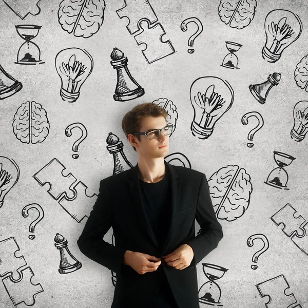 Nachdenklicher junger Geschäftsmann auf Betongrund mit kreativem Gehirn, Glühbirne, Fragezeichen und Schachfiguren-Zeichnungen. Lösungskonzept — Stockfoto