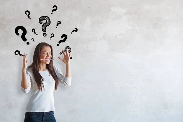 Portrait of furious young woman with pencil in mouth and hands up standing on concrete background with drawn question mark. Confusion concept — Stock Photo, Image