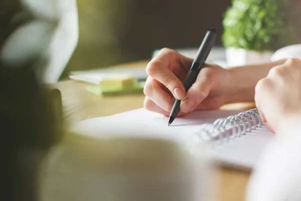 Primer plano de las manos de la niña escribiendo en espiral bloc de notas colocado en el escritorio de madera con elementos borrosos. Concepto educativo — Foto de Stock