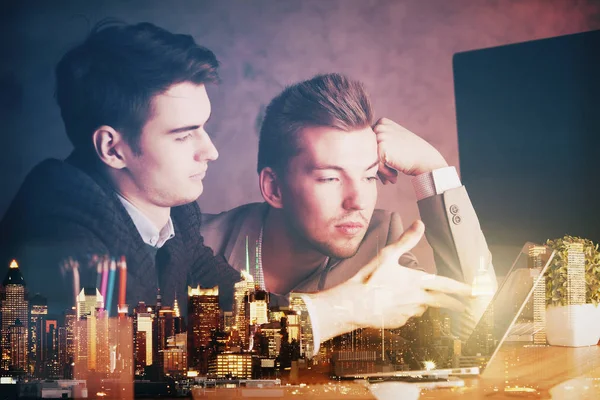 Zwei gutaussehende junge Männer, die Laptop benutzen und am Arbeitsplatz mit abstrakten nächtlichen Stadtansichten über ein neues Projekt diskutieren. Teamwork. Doppelbelastung — Stockfoto