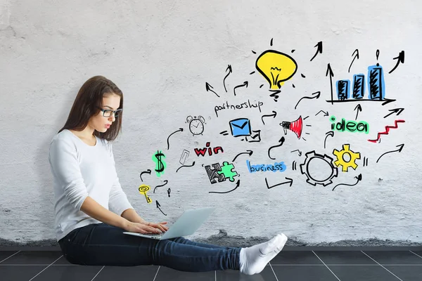 Seitenansicht von hübschen kaukasischen Mädchen auf dem Boden sitzend und mit Laptop in Betonraum mit bunten Business-Skizze an der Wand. Ideenkonzept — Stockfoto