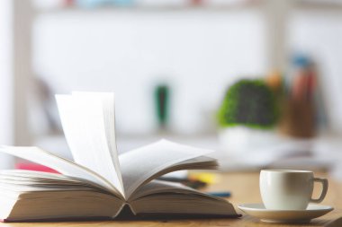 Ahşap ofis masaüstü seramik kahve fincanı, açık kitap, bulanık bitki, sarf malzemeleri ve diğer öğeleri ile kapatın. Eğitim kavramı