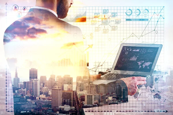 Young businessman using laptop on city background with business charts and graphs. Technology concept. Double exposure — Stock Photo, Image