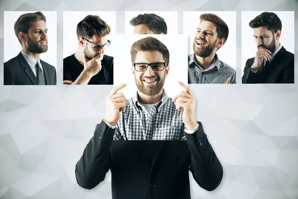 Junger europäischer Mann versteckt sich mit lächelndem Gesicht hinter einem Plakat. Reihe von Gesichtern mit unterschiedlichen Ausdrücken im Hintergrund. Emotionales Konzept — Stockfoto