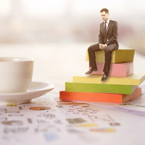Tiny thoughtful young businessman sitting on stickers placed on office desk with coffee cup and business sketch. Brainstorming concept — Stock Photo, Image