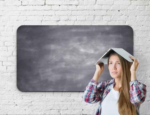 Bezorgd jonge vrouw staande naast leeg schoolbord en die betrekking hebben op kop met boek. Witte bakstenen muur achtergrond. Verwarring concept. Mock up — Stockfoto