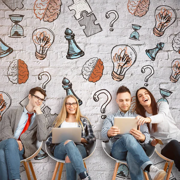 Cheerful young team sitting on chairs and using laptops on concrete background with creative drawings. Brainstorming concept — Stock Photo, Image