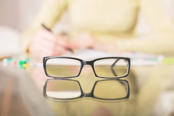 Primer plano de los espectáculos colocados en la mesa de cristal. Mujer borrosa en el fondo escribiendo en bloc de notas. Concepto de visión — Foto de Stock