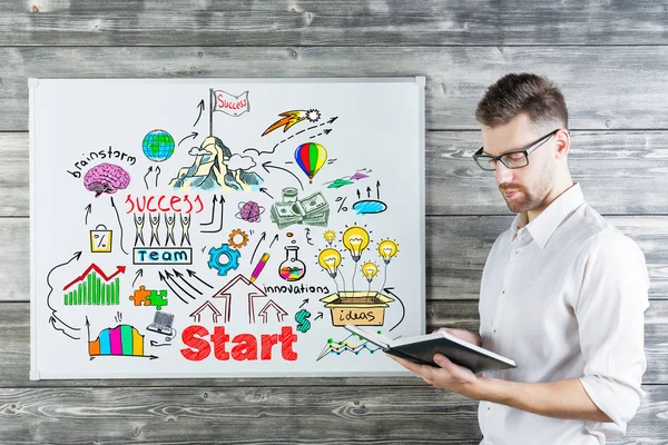 Aantrekkelijke jonge zakenman lezen boek naast whiteboard met zakelijke schets. Team concept. Houten muur achtergrond — Stockfoto