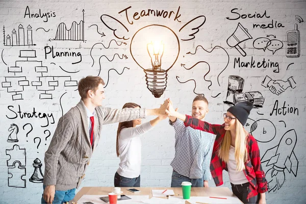 Unga europeiska team medlemmar Hej-fiving varandra över trä skrivbord. Tegelvägg med business skiss i bakgrunden. Lagarbete koncept — Stockfoto