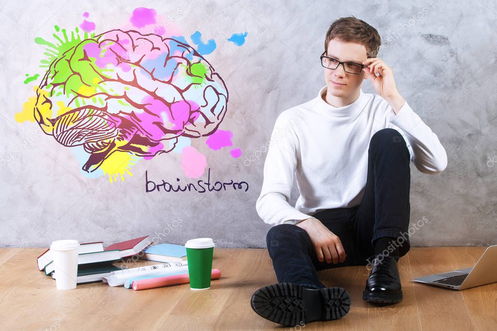 Young businessman sitting on floor with coffee cups, books, laptop and brain sketch on concrete wall. Brainstorm concept