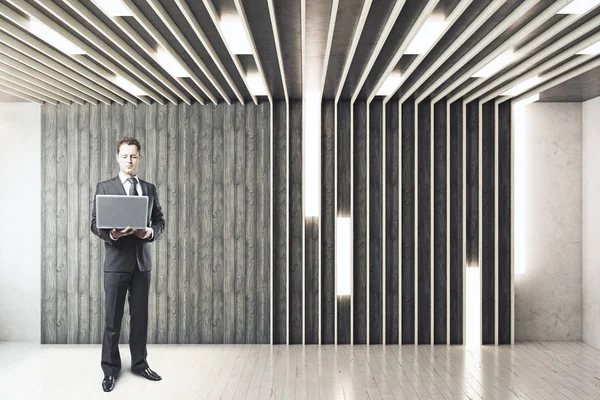 Pohledný mladý podnikatel s notebookem v ruce stojící v abstraktní interiéru. Komunikační koncept. — Stock fotografie