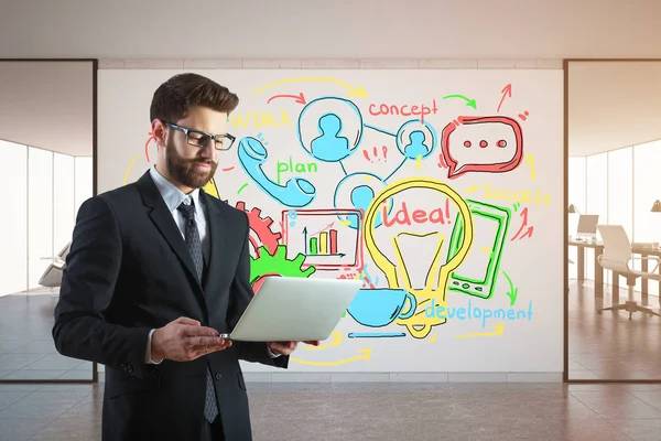 Handsome young businessman using laptop in modern office interior with colorful business sketch on billboard. Development concept. — Stock Photo, Image