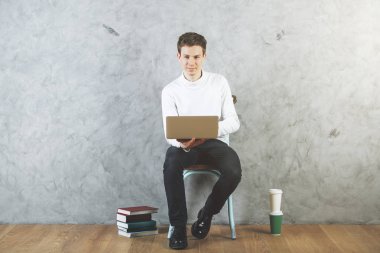 Yakışıklı genç adam sandalyede oturan ve beton duvar ile iç ve ahşap zemin kahve fincanı ve kitaplar ile dizüstü bilgisayar kullanarak. Öğrenci kavramı