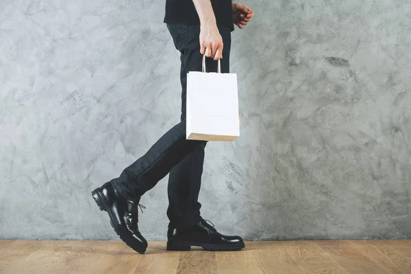 Vista lateral de las piernas del hombre y bolsa de compras vacía en las manos en la habitación con pared de hormigón y suelo de madera. Concepto de publicidad. Prepárate. —  Fotos de Stock