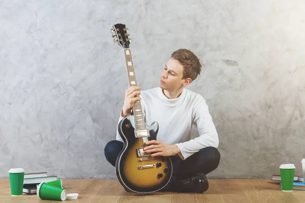 Schöner junger Mann spielt Gitarre, während er mit mehreren Kaffeetassen auf dem Holzboden sitzt. — Stockfoto