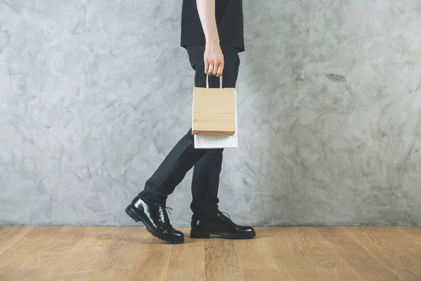 Vista lateral de las piernas del hombre y bolsa de compras vacía en las manos en la habitación con pared de hormigón y suelo de madera. Concepto de regalo. Prepárate. —  Fotos de Stock