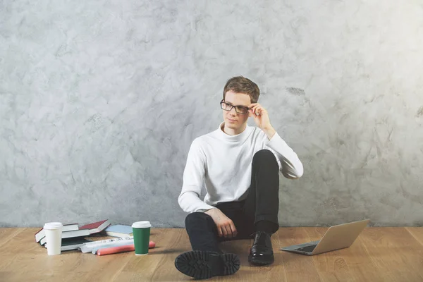 Gondolkodó fiatal üzletember ül a fa padló kávéscsészét, laptop, könyvek és más elem, beton alapon. Vizsga koncepció — Stock Fotó