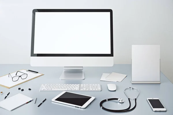 Gros plan du lieu de travail du médecin avec écran d'ordinateur blanc vide, autres appareils, paperasse et stéthoscope . — Photo