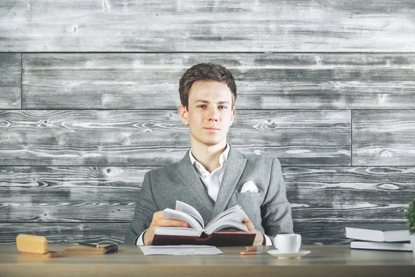 Handsome european businessman with book sitting at wooden desk with coffee cup and other items. Knowledge concept — Stock Photo, Image