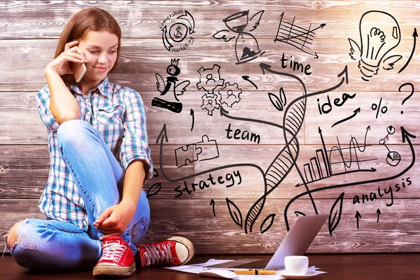 Glimlachen van jonge Europese vrouw zittend op houten vloer, praten over de telefoon en laptop scherm kijken met koffiekopje, schets papierwerk en zakelijke op muur. Leiderschap concept — Stockfoto