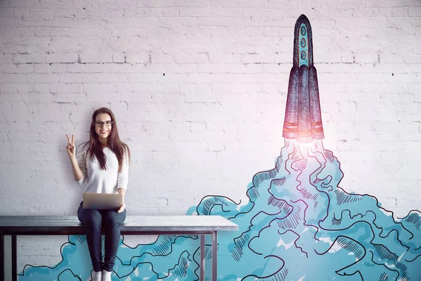 Mujer europea joven feliz sentado en la mesa con el ordenador portátil y mostrando el signo de la paz en el fondo de ladrillo blanco con el lanzamiento de boceto cohete. Concepto de emprendimiento — Foto de Stock