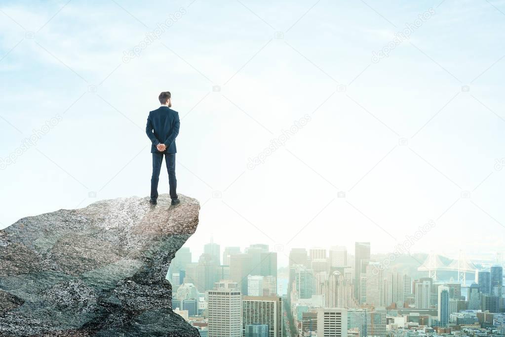 Back view of young businessman on mountain top looking at city with daylight. Future concept. Copy space