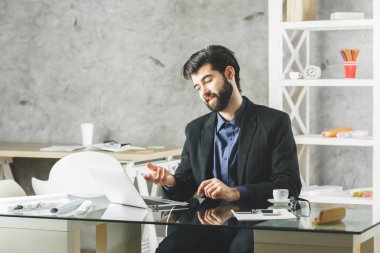 Modern ofis dizüstü bilgisayar ekranında cam masanın üzerinde çeşitli öğeleri ile bakan genç adam gülümsüyor. Beton duvar arka plan. İletişim kavramı 