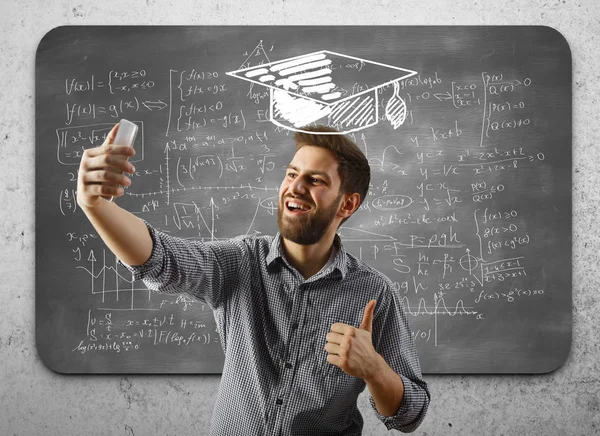 Vrolijke jonge zakenman met getekende mortarboard selfie te nemen en duimen opdagen op betonnen muur achtergrond met wiskundige formules op schoolbord. Succes concept — Stockfoto