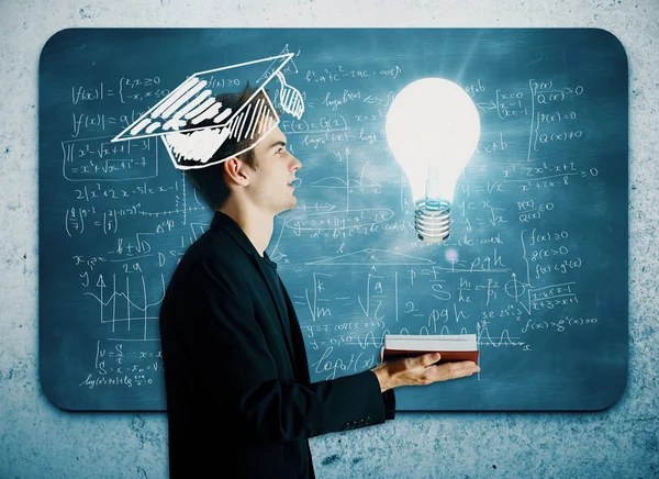 Handsome young businessman with drawn mortarboard holding book with glowing lamp on concrete wall background with mathematical formulas on chalkboard. Graduation concept — Stock Photo, Image