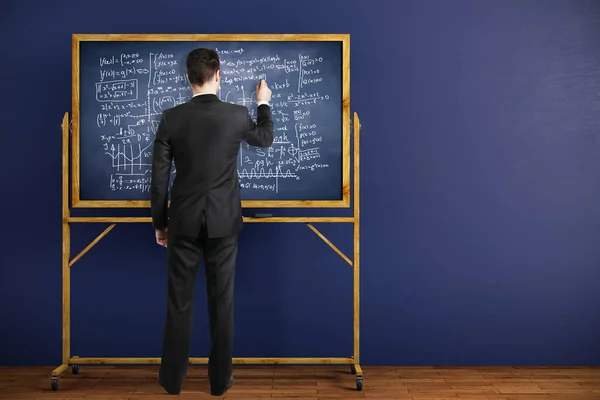 Achteraanzicht van zakenman schrijven van wiskundige formules op schoolbord geplaatst in interieur met blauwe muur en houten vloer. Onderwijs concept. 3D-rendering — Stockfoto