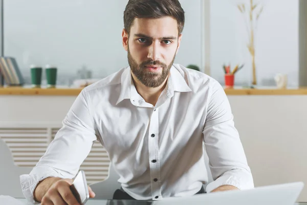 Retrato de un joven hombre de negocios europeo con smartphone en la mano usando el portátil en el escritorio de la oficina moderna. Concepto de trabajo —  Fotos de Stock