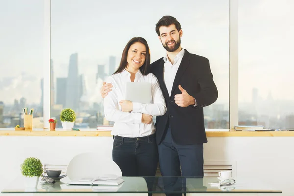 Beau homme et jolie femme montrant pouces vers le haut dans le bureau moderne avec vue floue sur la ville et bureau avec des articles. Concept réussi — Photo