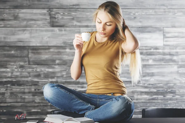 Atractiva mujer europea sentada en un escritorio de madera con taza de café y libro de lectura. Concepto de conocimiento — Foto de Stock