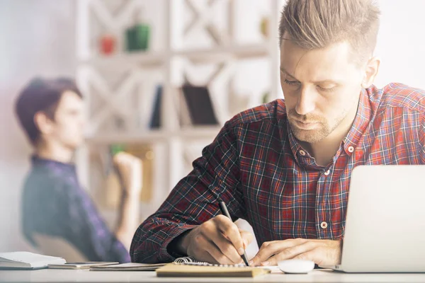 Retrato de menino branco bonito escrevendo em bloco de notas em espiral no local de trabalho. Conceito de papelada. Colega desfocada em segundo plano — Fotografia de Stock