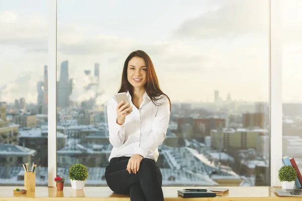 Atractiva joven sonriente usando un teléfono inteligente mientras está sentada en el alféizar de la ventana con artículos en el interior de la oficina con vistas borrosas a la ciudad. Concepto de comunicación — Foto de Stock