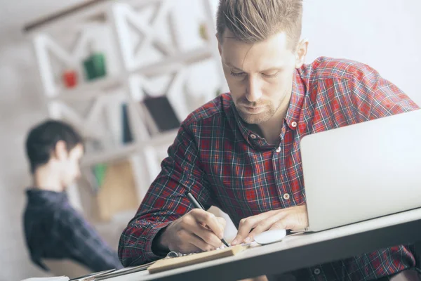 Retrato de la escritura masculina caucásica guapo en cuaderno en espiral en el lugar de trabajo. Concepto de papeleo. Colega borrosa en el fondo — Foto de Stock