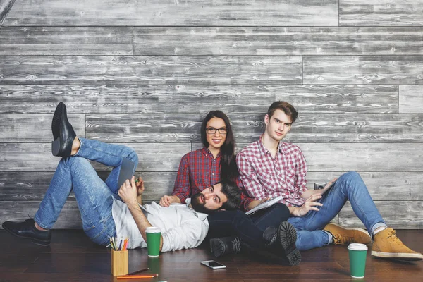 Atractivos hombres de negocios y mujeres con taza de café, suministros y dispositivos sentados en el piso del interior del estudio de madera. Concepto divertido —  Fotos de Stock