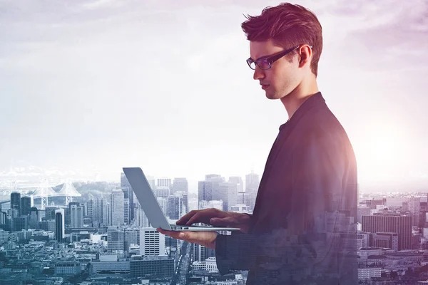 Vista laterale del giovane imprenditore europeo che utilizza laptop su sfondo astratto della città con luce del giorno. Il concetto di comunicazione. Doppia esposizione — Foto Stock