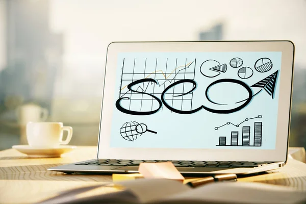 Close up of creative office desktop with SEO sketch on laptop screen, coffee cup and other items on blurry city background. Web concept — Stock Photo, Image