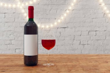 Boş etiket ve cam ahşap yüzey ve tuğla duvar arka plan üzerinde kırmızı şarap şişe dekoratif lambalar ile yakın çekim. Kadar alay et. 3D render