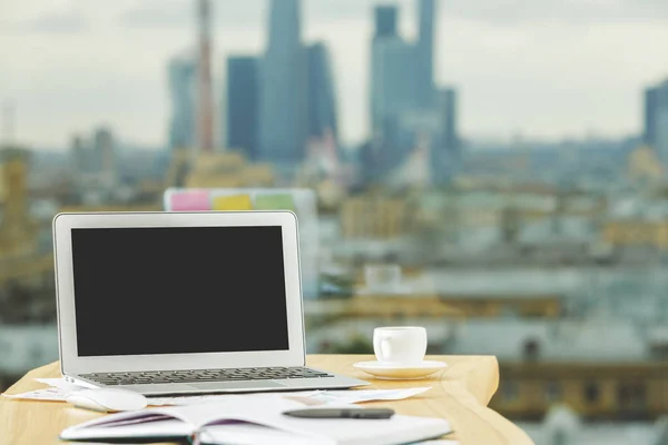 Close up of creative table with empty laptop, coffee cup and supplies on blurry city view background. Mock up
