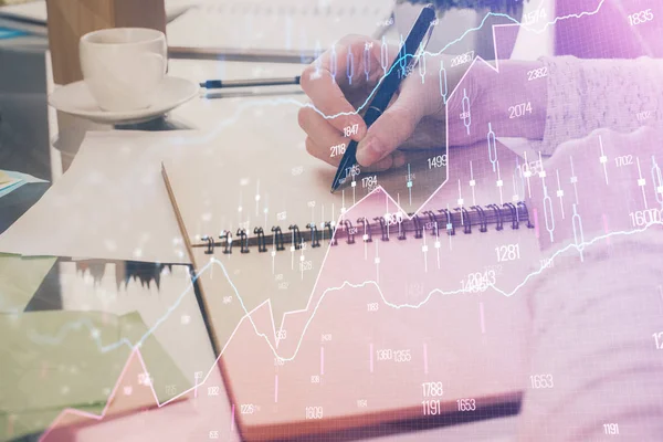 Hand taking notes in notepad. Forex chart holograms in front. Concept of research. Double exposure