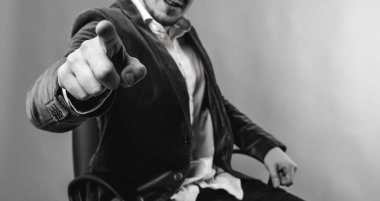 İş adamı, katı ve iş adamı, kahverengi ceketli ve beyaz gömlekli, bir sandalyeye oturur ve parmağını önde, mavi arka planda gösterir.