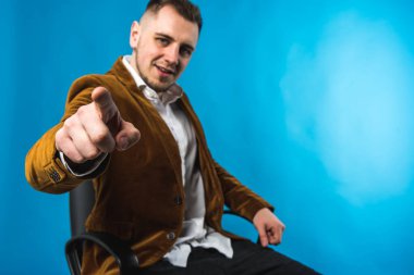 İş adamı, katı ve iş adamı, kahverengi ceketli ve beyaz gömlekli, bir sandalyeye oturur ve parmağını önde, mavi arka planda gösterir.