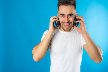 Beyaz tişörtlü yakışıklı Avrupalı adam müzik dinliyor, mavi arka planda büyük siyah kulaklıklar, duygusal. 