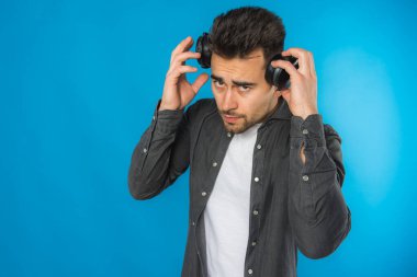 Gri tişörtlü ve beyaz tişörtlü yakışıklı Avrupalı genç müzik dinliyor, mavi arka planda büyük siyah kulaklıklar, duygusal, farklı el hareketleri sergiliyor.