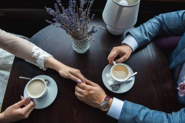 Damat gelinin elini avucunun içinde tutar gelinin buketinin arka planında bir masada otururlar ve yakın plan kahve içerler.